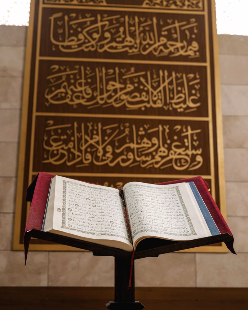 An open Quran on a wooden stand beneath mosque calligraphy