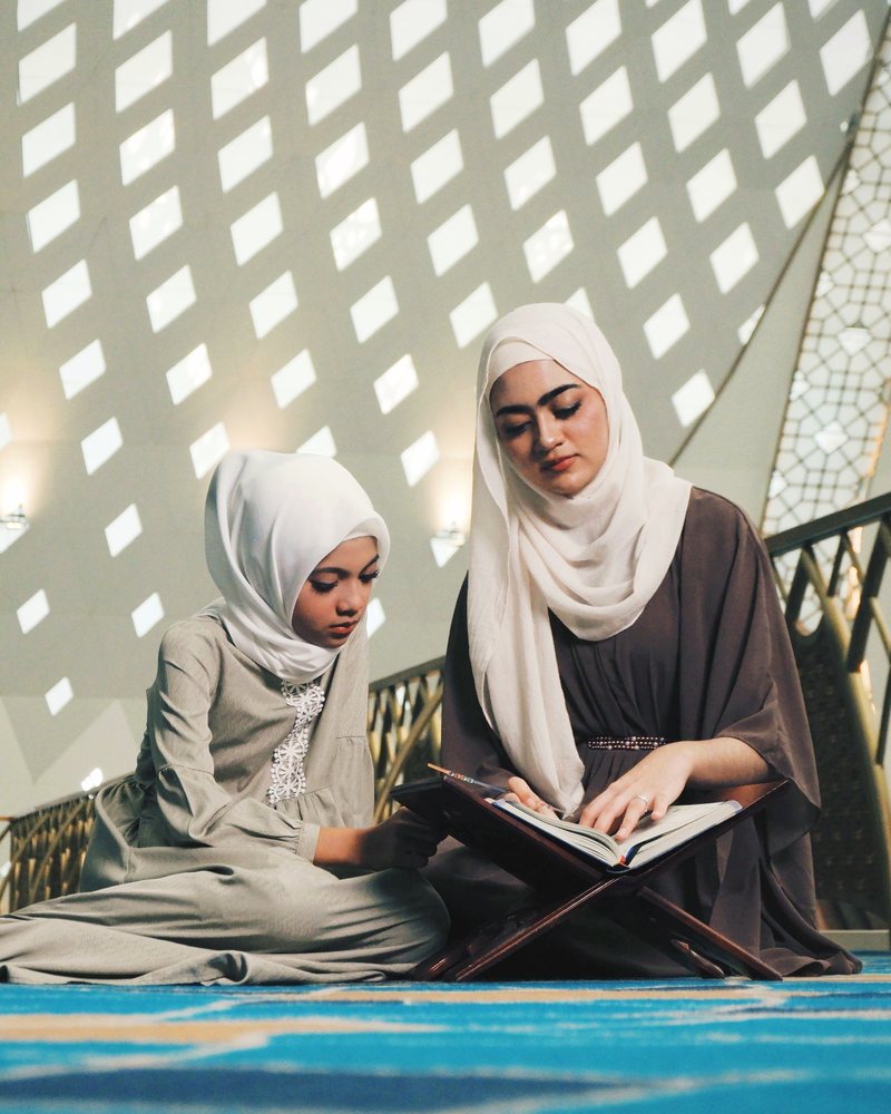 A mother in a hijab helping her child read the Quran on a wooden book stand