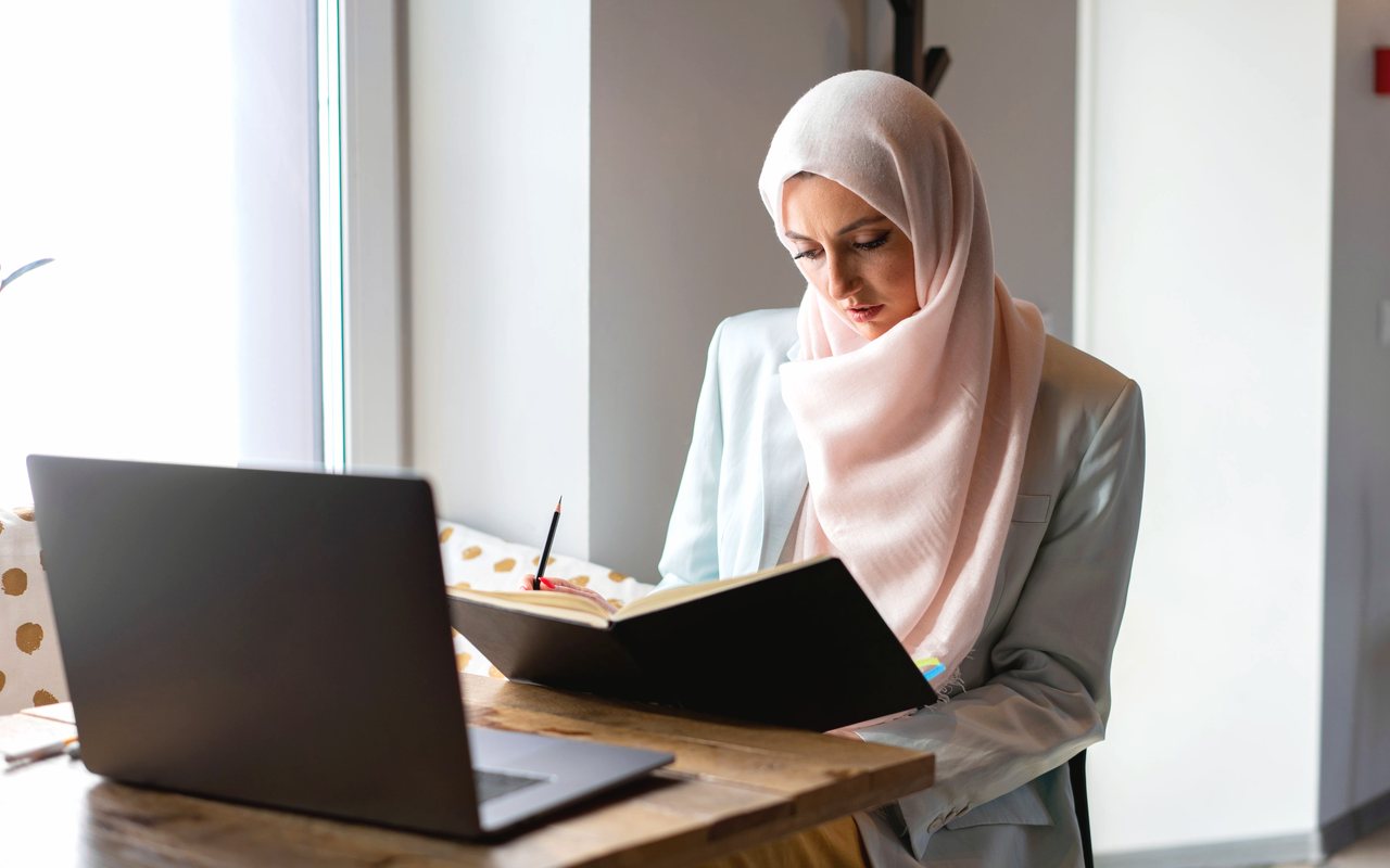 A woman in a hijab following a live online lesson with a book and a laptop